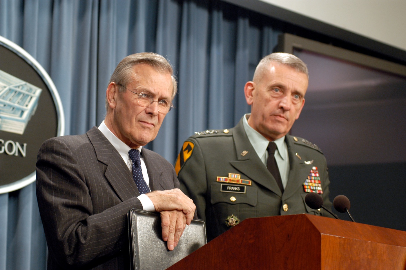 Two men at a Pentagon press briefing podium, one in a suit and one in an Army uniform.